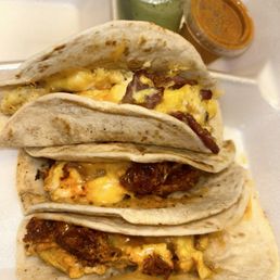 Mixed Breakfast Taco - Three breakfast tacos with flour tortillas filled with scrambled eggs, melted cheese, and meat, possibly bacon or sausage. The tacos are served in a foam container with small cups of green and red salsa or sauces in the background. The image highlights traditional Mexican breakfast offerings.