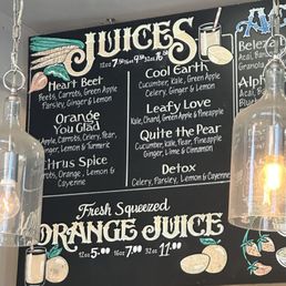 Menu May 2025 - The image shows a black chalkboard menu with colorful hand-drawn illustrations of fruits and a glass of juice. The menu lists various organic juice options such as Heart Beet, Orange You Glad, Citrus Spice, Cool Earth, Leafy Love, Quite the Pear, and Detox, with their ingredients. The menu also features prices for different sizes (12 oz, 16 oz, 32 oz). Two glass pendant lights hang on either side of the chalkboard, illuminating the area.