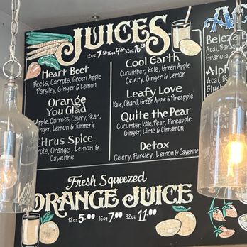 Main Squeeze - Menu May 2025 - The image shows a black chalkboard menu with colorful hand-drawn illustrations of fruits and a glass of juice. The menu lists various organic juice options such as Heart Beet, Orange You Glad, Citrus Spice, Cool Earth, Leafy Love, Quite the Pear, and Detox, with their ingredients. The menu also features prices for different sizes (12 oz, 16 oz, 32 oz). Two glass pendant lights hang on either side of the chalkboard, illuminating the area. - Juice Bars & Smoothies Near Me - Oakland, California