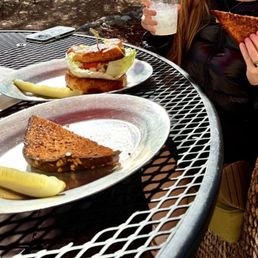 Grilled cheese and chicken salad sandwich! (And lemonade) - The image shows an outdoor metal table with two plates of food, one with a slice of cake and a lemon wedge, and the other with a sandwich. A person, partially visible, holds a grilled cheese sandwich in one hand and a drink in the other. The setting appears to be a patio or garden area, with natural lighting and some background foliage.