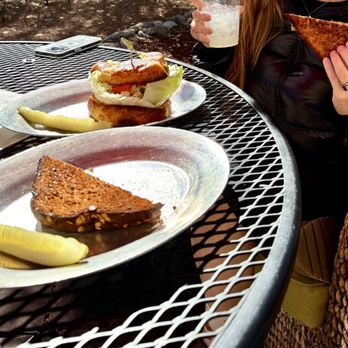 Cafe Canyon - Grilled cheese and chicken salad sandwich! (And lemonade) - The image shows an outdoor metal table with two plates of food, one with a slice of cake and a lemon wedge, and the other with a sandwich. A person, partially visible, holds a grilled cheese sandwich in one hand and a drink in the other. The setting appears to be a patio or garden area, with natural lighting and some background foliage. - Coffee & Tea Near Me - Santa Fe, New Mexico