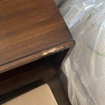 Apache Moving & Storage - Damaged wood table - A wooden furniture item shows visible damage on its edge, with chipped and worn areas, positioned near a plastic-covered mattress. - Movers Near Me - Leander, Texas