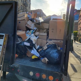 GreenEx CleanOut Services - A truck parked on a city sidewalk is filled with a large pile of cardboard boxes, plastic bags, and miscellaneous waste. The background features multi-story buildings, a bright sky with the sun shining, and pedestrians walking along the sidewalk. The scene suggests a cleanup or waste disposal activity in an urban environment. - Junk Removal & Hauling Near Me - Flushing, New York