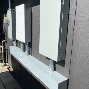 Comletric - Multiple electrical enclosures and conduit pipes are installed on an exterior wall, with a metal cable tray beneath them, against a dark siding background. - Electricians Near Me - Concord, California