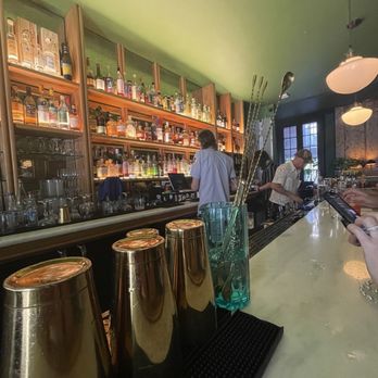 Peychaud’s - Light wooden varnished liquor shelving underneath the absinthe green ceiling with polished granite counter tops and 30s ceiling lighting. - Cocktail Bars Near Me - New Orleans, Louisiana Peychaud’s - Light wooden varnished liquor shelving underneath the absinthe green ceiling with polished granite counter tops and 30s ceiling lighting. - Cocktail Bars Near Me - New Orleans, Louisiana