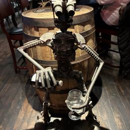 A robot sculpture made of metal parts is positioned in front of a wooden barrel in a restaurant, holding a wine glass.