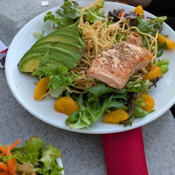 RA Sushi - Mesa - Nutty grilled salad with salmon - A white plate contains a salad with grilled salmon, sliced avocado, mixed greens, and yellow fruit or vegetable pieces, along with noodles. The plate is on a gray table, with chopsticks held by a person in the background. Additional salad is visible in a bowl in the foreground. The setting appears to be an outdoor dining area. - Japanese Near Me - Mesa, Arizona