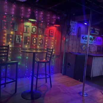 Nu Towne Saloon - The image shows a bar interior with a small stage area that has two high chairs and a round table. The background is decorated with multicolored string lights and a sign that reads 'WELCOME BALCON.' Various signs, including traffic signals and business advertisements, are mounted on the wooden wall. The lighting is dim with colorful accents, creating a lively atmosphere suitable for social gatherings. - Gay Bars Near Me - Phoenix, Arizona