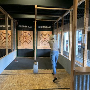 Oregon Axe - Working to throw the axe correctly - A woman with blonde hair, wearing a beige top and blue jeans, is in the process of throwing an axe at a target in an indoor axe throwing facility. - Axe Throwing Near Me - Springfield, Oregon