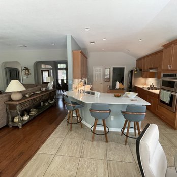 Eagle Design Build - A kitchen features a white island with bar stools, wooden cabinets, and stainless steel appliances, connected to a living area with a sofa. - General Contractors Near Me