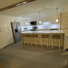 A spacious kitchen with white cabinets and marble countertops. The kitchen includes a marble island with four beige stools, stainless steel refrigerator, microwave, and oven. Pendant lights hang above the island, and the space has a neutral color palette with light wood flooring.
