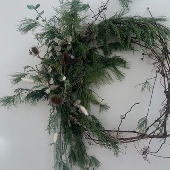 Constellation Coffee - A floral arrangement composed of various green foliage, including pine and eucalyptus, with dried elements such as seed pods and cotton-like fibers. The arrangement is laid out on a plain white background, with some branches extending outward and ribbons hanging from the bottom. - Coffee & Tea Near Me - Pittsburgh, Pennsylvania