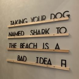 a sign that says taking your dog named shark to the beach is a bad idea - The image shows a decorative wall sign with black letters attached to horizontal wooden slats. The sign displays a humorous quote about dogs and the beach, reading: 'TAKING YOUR DOG NAMED SHARK TO THE BEACH IS A BAD IDEA.' The sign is mounted on a plain, light-colored wall, and the letters are arranged in four lines with spacing between the slats.