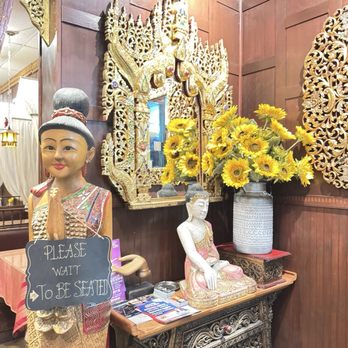 Ploy Thai - The image shows a corner of a restaurant interior decorated with Thai-style statues, including a standing figure with a sign that reads 'Please wait to be seated.' There are ornate gold carvings and a mirror framed with intricate gold designs. A large vase filled with yellow sunflowers is placed on a small table, alongside brochures and decorative items. The wood-paneled wall adds warmth to the scene. - Thai Near Me - Brandon, Florida