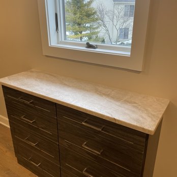River Road Tops - Taj Mahal quartzite, leather finish in closet - A dark wood cabinet featuring several drawers with metallic handles is situated beneath a window. The cabinet has a light-colored marble top. The window provides a view of a neighboring house and trees outside, with some snow visible on the ground. The room has hardwood flooring and neutral-colored walls. - Cabinetry Near Me - Lockport, Illinois