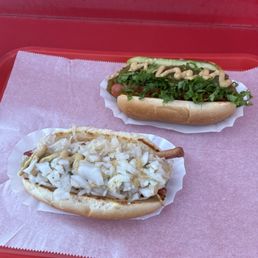 Two hot dogs with different toppings are served on a pink tray with pink paper, one topped with chopped onions and the other with lettuce, tomatoes, and sauce.
