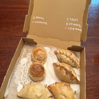 Che Empanada - Creamy Chicken Empanada,  Ham & Cheese Empanada,  Beef Criolla Empanada, and a short rib empanada. - The image shows an open cardboard box with six baked empanadas and pastries arranged inside, lined with paper doilies. The box is placed on a wooden table, and the interior of the lid has handwritten notes indicating the contents, such as '2 empanadas' and '1 shot of cream.' The empanadas vary in shape and size, with some having decorative crimped edges and others with small protrusions, indicating different fillings or styles. - Empanadas Near Me - Worcester, Massachusetts