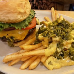 Green chili cheese burger with green chili cheese fries. - A cheeseburger features a beef patty topped with melted cheese, lettuce, and tomato, served in a bun. It is accompanied by a side of green chili cheese fries, which are covered with melted cheese and chopped green chilies. The meal is presented on a white plate, likely at a casual restaurant setting.