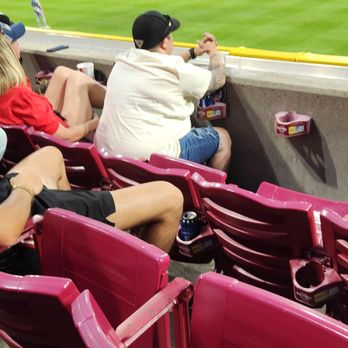 Cincinnati Reds - People are seated in stadium chairs at a baseball game, facing the field. The image shows a view of the outfield, with players on the field and the stadium's architecture in the background. The seats are pink, and some spectators have drinks and personal items nearby. The stadium is well-lit, indicating an evening game, with a large crowd present. - Professional Sports Teams Near Me - Cincinnati, Ohio