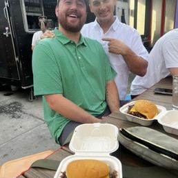 Burger - Two men are seated at an outdoor table, smiling at the camera. The man on the left wears a green and white striped polo shirt and a cap, while the man on the right wears a white button-up shirt and points at his friend. On the table, there are takeout containers with burgers, one with a beef patty, cheese, and a bun. The background includes a food truck and a building with colorful murals, indicating a casual outdoor dining setting.