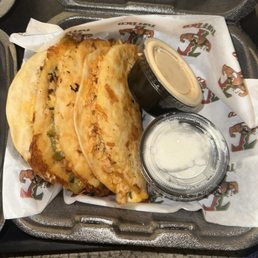 Chicken sexy tacos - Three tacos with various fillings are placed on a tray lined with branded paper, accompanied by two small containers of sauces.