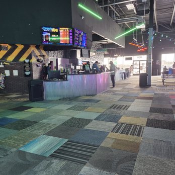 Combat Ops - The image shows a large indoor entertainment facility with a concession counter equipped with digital menu screens displaying various options. The space includes seating areas with tables and chairs, arcade game machines, and a high ceiling with exposed beams and colorful lighting fixtures. Natural light enters through large windows at the far end, and a few people are visible near the counter and seating area, indicating a lively environment. - Laser Tag Near Me - Indianapolis, Indiana