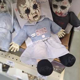 Unbelievable, it's the middle of August! They put this crap in a main location in both stores. - The image features a horror-themed doll with a cracked, zombie-like face, dark hollow eyes, and a distressed expression. It is dressed in a blue dress with a white collar and has a tag that says "TRY!" on its chest. The doll is seated on a store shelf among other similar creepy dolls, with visible price tags indicating it is for sale. The overall scene suggests a display of Halloween or horror collectibles.