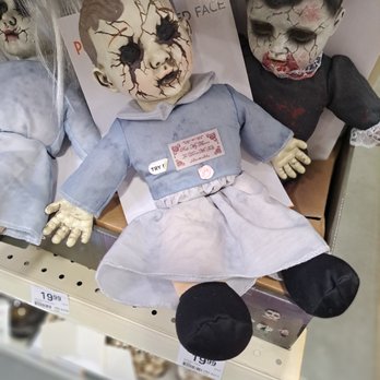 Menards - Unbelievable, it's the middle of August! They put this crap in a main location in both stores. - The image features a horror-themed doll with a cracked, zombie-like face, dark hollow eyes, and a distressed expression. It is dressed in a blue dress with a white collar and has a tag that says "TRY!" on its chest. The doll is seated on a store shelf among other similar creepy dolls, with visible price tags indicating it is for sale. The overall scene suggests a display of Halloween or horror collectibles. - Building Supplies Near Me - Kalamazoo, Michigan