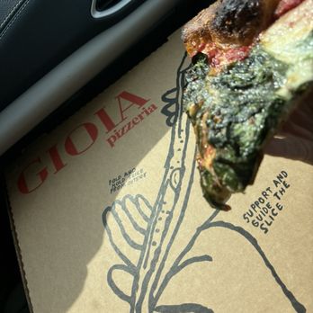 Gioia Pizzeria - A hand holds a slice of pizza with spinach, cheese, and a slightly burnt crust inside a vehicle. The pizza is positioned over a Gioia Pizzeria box, which features a simple illustration of a hand holding a pizza slice and instructions for folding and supporting the slice. The interior of the car includes a black dashboard and air vent. - Pizza Near Me - Berkeley, California