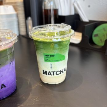 Matcha Matcha - Tokyo Fog - A layered matcha latte in a clear plastic cup displays green tea and milk layers, with a green label reading 'mat-cha' and 'MATCHA.' The cup has a plastic lid and is placed on a black countertop, with a blurred background including other beverage containers and a black storage tray. - Coffee & Tea Near Me - Markham, Ontario