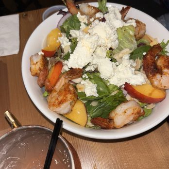 Tapped - A salad featuring cooked shrimp, fresh mixed greens, sliced peaches, and crumbled cheese is served on a white bowl. The salad includes vegetables such as carrots and possibly onions, with a variety of textures and colors. A drink with ice and a straw is placed in front of the plate on a wooden table. The setting appears to be a casual dining environment. - Cocktail Bars Near Me - Middletown, New York