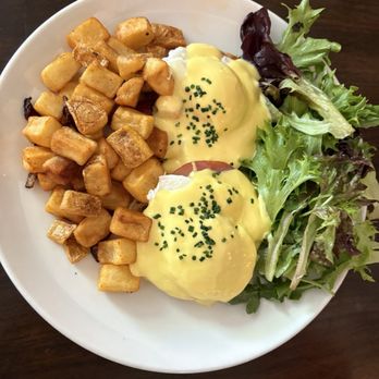 Agustin Kitchen - Eggs Benedict - A plate features Eggs Benedict topped with hollandaise sauce and chives, served with breakfast potatoes and a side of mixed greens. - New American Near Me - Tucson, Arizona