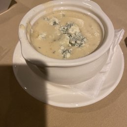 Blue Cheese Soup - A ceramic bowl filled with blue cheese soup, topped with crumbles of blue cheese, placed on a white plate with a napkin, on a beige table.
