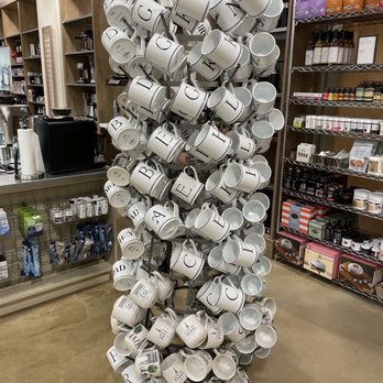 Sur La Table - The image shows a retail store interior with a metal stand holding many white coffee mugs with black lettering, arranged in a spiral pattern. The store has wooden shelving units stocked with various products, including packaged food items and kitchen accessories. The floor is a polished concrete surface, and the background includes a counter with a coffee machine and other store items. - Kitchen Supplies Near Me - Chicago, Illinois