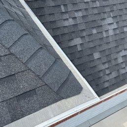 The photograph captures a section of a building's roof, featuring two different types of asphalt shingles: one with a granular texture and another with a smoother, darker surface. A metal gutter runs along the edge of the roof, with visible fasteners. Below the roofline, part of a brick wall and a concrete surface are visible, indicating the building's exterior. The image provides a clear view of roofing materials and gutter installation.