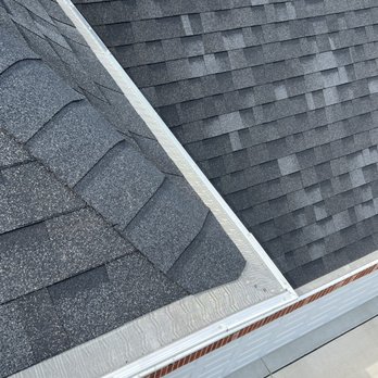 Ultimate Gutter - The photograph captures a section of a building's roof, featuring two different types of asphalt shingles: one with a granular texture and another with a smoother, darker surface. A metal gutter runs along the edge of the roof, with visible fasteners. Below the roofline, part of a brick wall and a concrete surface are visible, indicating the building's exterior. The image provides a clear view of roofing materials and gutter installation. - Gutter Services Near Me - Schaumburg, Illinois