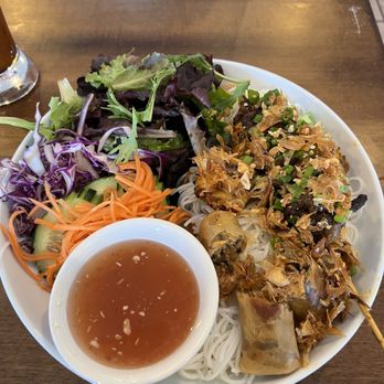 Bamboo by Kulantro - A plate of Vietnamese cuisine includes spring rolls with shredded meat and vegetables, rice noodles topped with fried shallots and chopped green onions, and a side salad with mixed greens, shredded carrots, and cucumber. A small bowl of dipping sauce with garlic and chili flakes is also present. The dish is served on a white, round plate placed on a wooden table, with a glass of water and a glass of dark beverage visible in the background. - Vietnamese Near Me - Albuquerque, New Mexico