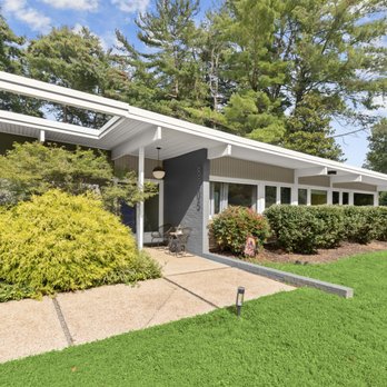 Badger Contracting - The image shows a mid-century modern style house with a flat, overhanging roof and extensive glass windows. The front yard features neatly trimmed bushes, a concrete pathway, and a lush green lawn. Tall trees are visible in the background, and the sky is partly cloudy, indicating a bright day. The house has a contemporary design with clean lines and a minimalist aesthetic. - Roofing Near Me - Pikesville, Maryland