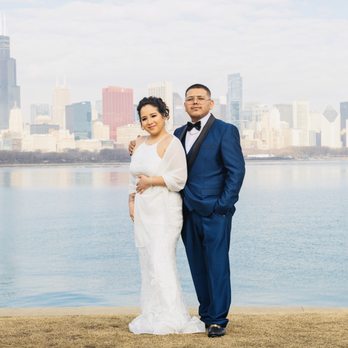 MNPhotoStudios - A bride and groom stand together on a grassy area beside a body of water, with a city skyline featuring tall buildings and skyscrapers in the background. The bride wears a white wedding gown with lace details, and the groom is dressed in a blue suit with a black bow tie. The scene appears to be a wedding or engagement photo shoot in an urban outdoor setting. - Session Photography Near Me - Chicago, Illinois