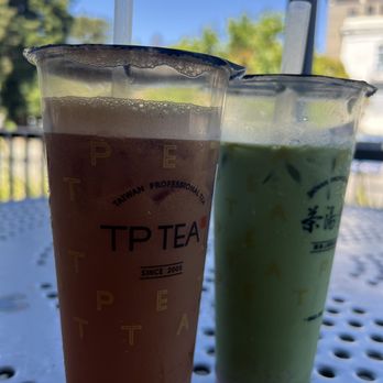 TP TEA - Two iced tea drinks are positioned on a perforated metal table outdoors. The left drink is brown, likely a traditional tea, and the right drink is green, possibly a matcha or green tea. Both drinks are in clear plastic cups with straws, and the cups have branding from TP Tea, a Taiwanese tea brand established in 2005. The background features a blue sky, trees, and a building, indicating a sunny day in an outdoor seating area. - Coffee & Tea Near Me - Berkeley, California