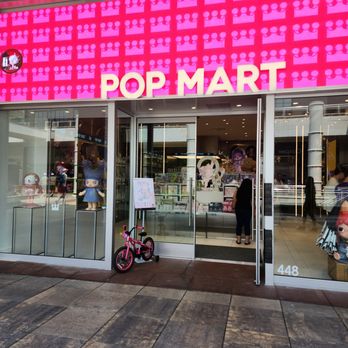 Pop Mart - The image shows the storefront of Pop Mart in Oak Brook, IL, with large glass windows revealing toys and collectible figures inside. The store has a pink sign with white letters spelling 'POP MART' above the entrance. A pink bicycle is parked outside on the sidewalk, and the store's address number 448 is visible on the right side of the window. The interior displays various toys, and a few customers are seen browsing inside. - Toy Stores Near Me - Oak Brook, Illinois