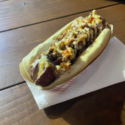 Ditty Beef Dog - A hotdog with a sausage in a bun is topped with melted cheese, caramelized onions, crunchy toppings, and drizzled with sauces. It is served in a paper tray on a wooden table, with a napkin underneath. The background shows a blurred surface, possibly part of a table or counter.