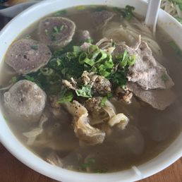Kulantro Pho - A large white bowl contains Vietnamese pho with slices of beef, meatballs, and chopped green onions on top. The broth is clear and filled with rice noodles. Fresh herbs and seasonings are visible, and a white spoon rests inside the bowl. The dish is served on a wooden table, with small side dishes and condiments in the background.