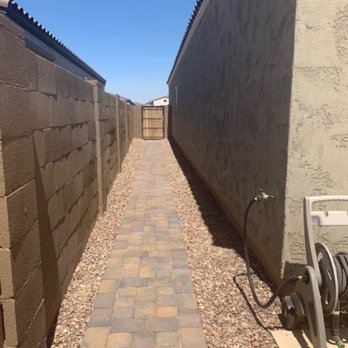 Diamond Cut Landscaping - walkway added from driveway extension - A narrow brick pathway, constructed with multicolored pavers, extends straight ahead between a dark brown brick wall on the left and a light-colored stucco wall on the right. Gravel lines both sides of the pathway, and a garden hose reel is visible on the right side near the foreground. The pathway leads to a wooden gate at the end, with a clear blue sky overhead, indicating a sunny day. - Landscaping Near Me - Phoenix, Arizona