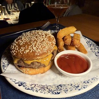 6 NYC - Burger - A plate features a cheeseburger with a sesame seed bun, melted cheese, and a beef patty. Next to the burger are onion rings and a small dish of ketchup. A glass of rosé wine is placed behind the plate. The setting appears to be a restaurant with other diners visible in the background, and the plate is on a dark blue placemat. - New American Near Me - Brooklyn, New York