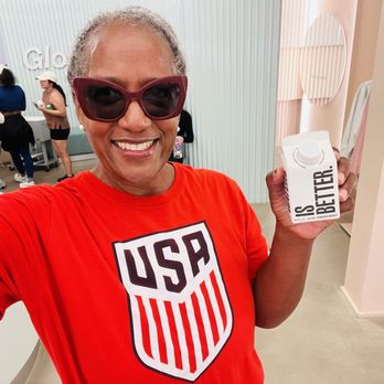 Glossier Chicago - Water refresher - A woman with gray hair and sunglasses is taking a selfie, holding a carton of water labeled 'Better.' She is in a modern store with other people in the background. - Cosmetics & Beauty Supply Near Me - Chicago, Illinois