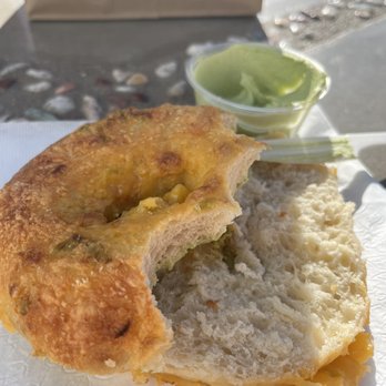 Sunday Bagels - A bagel with melted cheese and green spread in a small cup, set on a white paper plate outdoors. - Bagels Near Me - Albuquerque, New Mexico