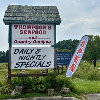 Thompson Seafood - The image shows a large outdoor signboard for Thompson's Seafood, featuring a shingled roof design at the top. The sign advertises seafood and country cooking, with a focus on daily and nightly specials. An 'Open' flag is positioned beside the sign, and a smaller blue sign provides a relief contact number for Hurricane Helene assistance. The setting includes greenery, wooden logs, and a paved area, indicating a rural or semi-rural location. - Seafood Near Me - Deep Gap, North Carolina