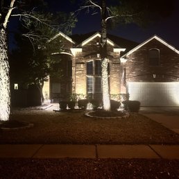 Landscape Lighting - 8 up lights and 5 path lights. All Kichler brand with brilliance LED bulbs. - The image shows a two-story brick house illuminated by outdoor landscape lighting, including up lights and path lights. The lighting accentuates the house's architectural features, trees, and bushes in the front yard. The scene is set at night, with a dark sky and shadows cast by the lighting. The driveway and sidewalk are visible in the foreground.