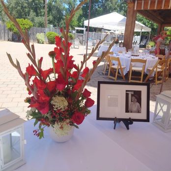 Planted on Main  - Celebration of life arrangement - A flower arrangement with red roses and gladiolus, accented with white filler flowers, is displayed on a table at an outdoor gathering with tables and chairs. - Florists Near Me - Placerville, California